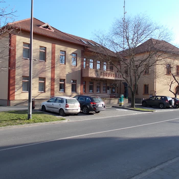 Monor városházának épülete, sárga-okkersárga vakolat, piros cseréptető, emeleti erkéllyel; előtte parkoló személyautók és kopasz fák, tiszta kék ég.